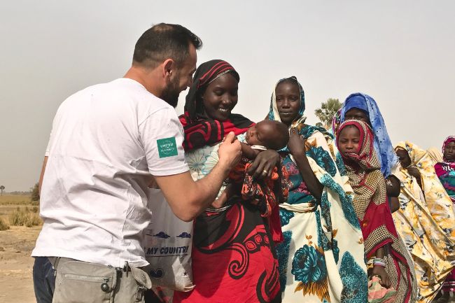 Ein humanitärer Helfer in Benin lächelt eine Mutter mit Baby auf dem Arm an und übergibt ihnen eine Tüte mit Kurban.