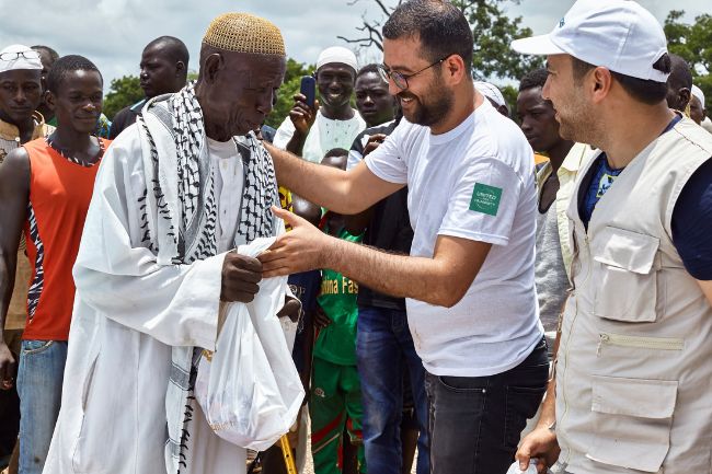 Ein älterer Mann in Burkina Faso erhält ein Kurban. Er ist umgeben von fröhlichen Hilfskräften und Begünstigten.